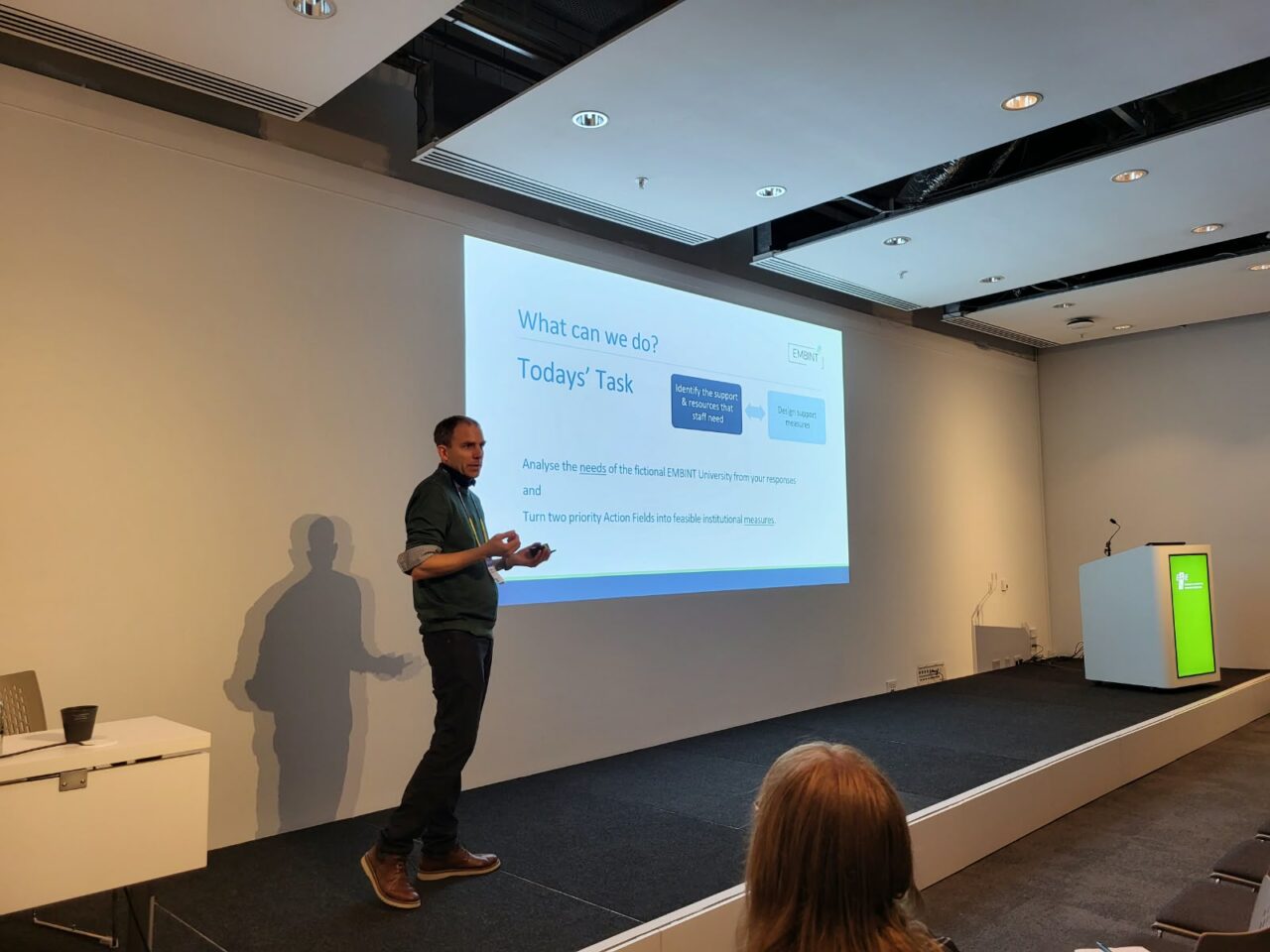 Johannes Weinreich presenting on a stage in a conference room. The projected slide behind him reads "What can we do? Today's Task" and asks participants to analyse the needs of a fictional "EMBINT University" and turn two priority action fields into feasible institutional measures. An audience member is visible in the foreground.