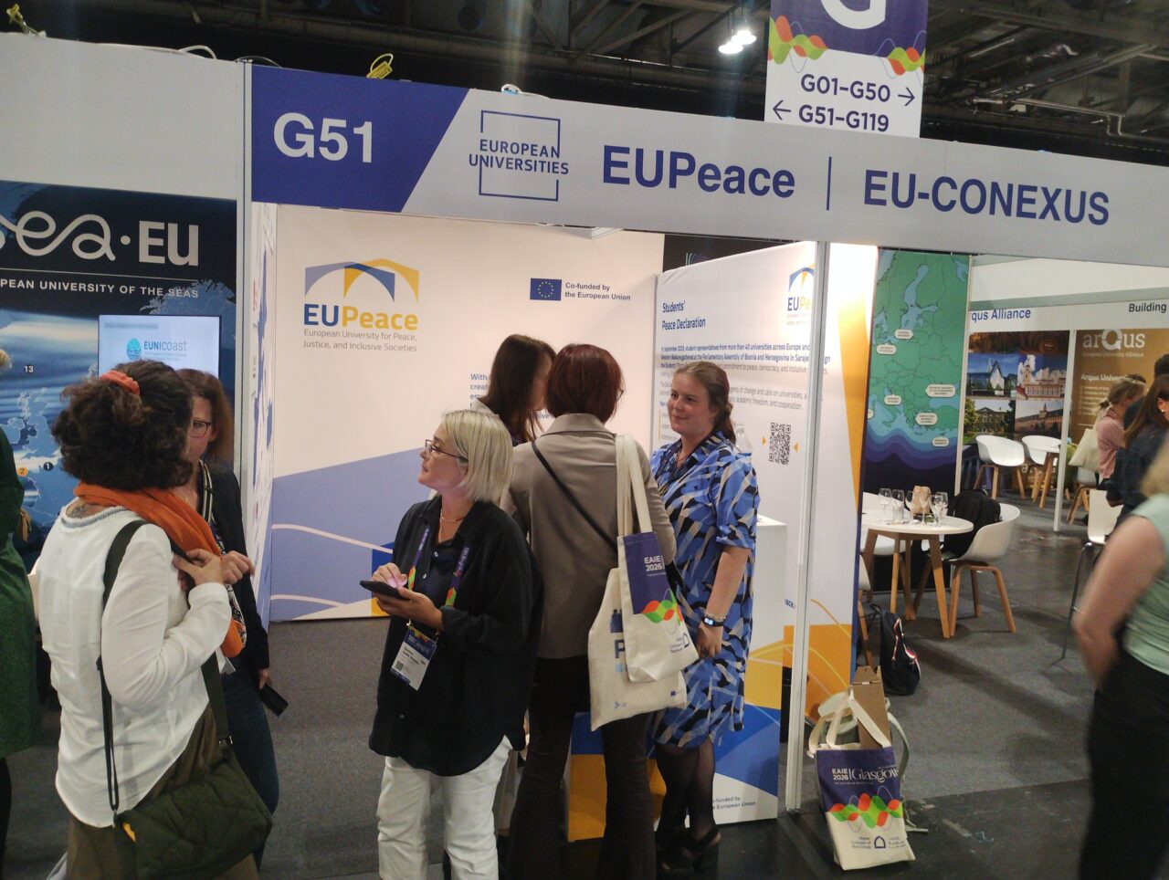 Conference visitors talking at a shared exhibition booth marked G51. The overhead banner reads "European Universities", "EUPeace" and "EU-CONEXUS". Behind the group are a EUPeace wall with the EUPeace logo and the "Co-funded by the European Union" emblem, and a roll-up banner titled "Students' Peace Declaration" with a QR code. Neighbouring stands of other European university alliances are visible on both sides.