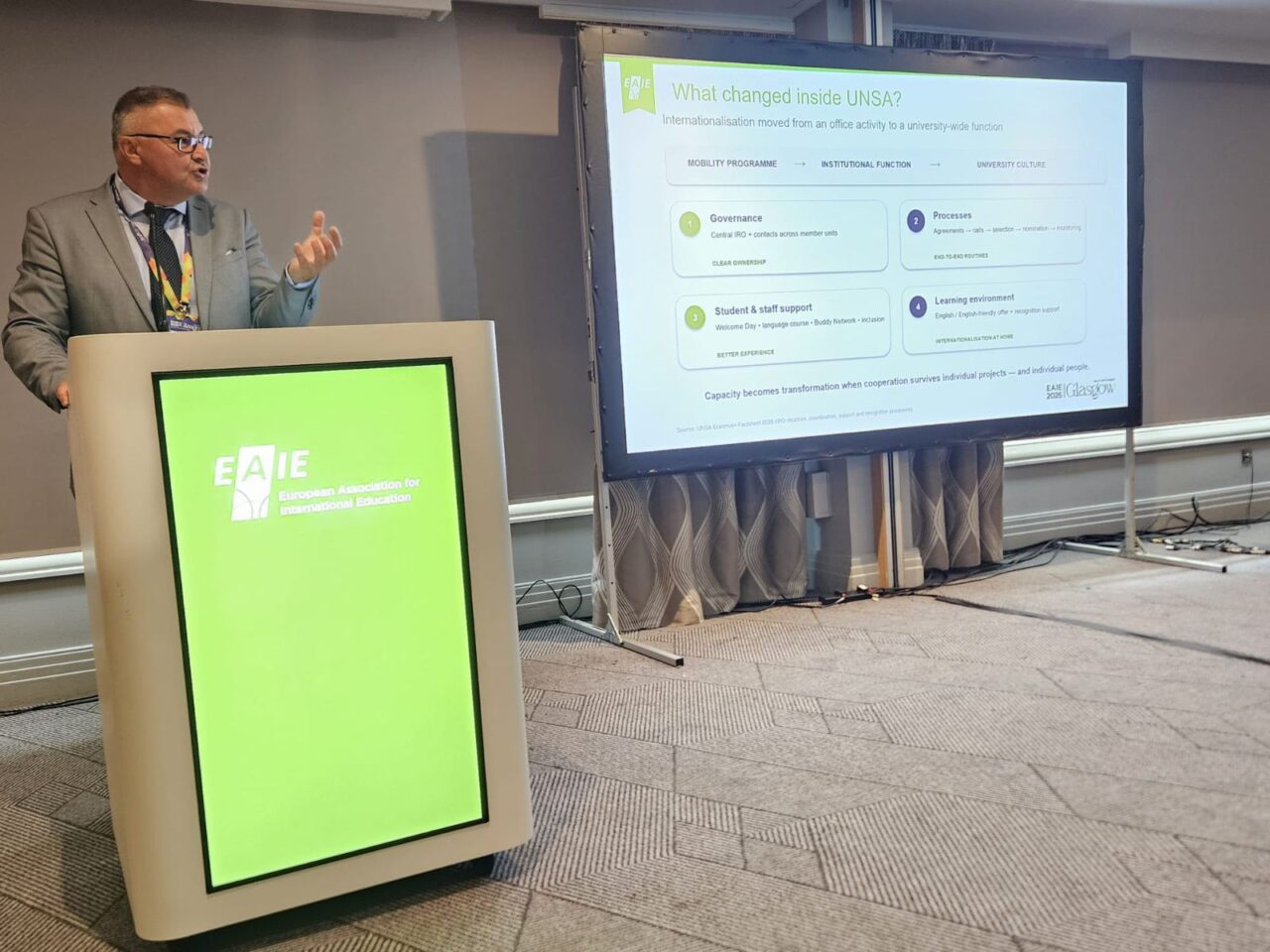 Kemal Durić speaking at a lectern with a green EAIE (European Association for International Education) display, gesturing with one hand. Beside him, a screen shows a slide titled "What changed inside UNSA?" with the subtitle "Internationalisation moved from an office activity to a university-wide function". The slide shows the progression "Mobility programme – Institutional function – University culture" and four numbered fields: Governance, Processes, Student & staff support, and Learning environment.