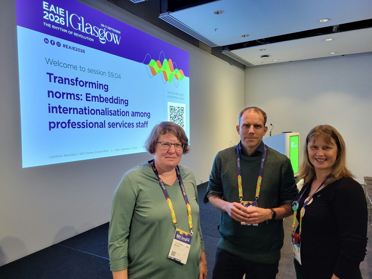 Christina Bohle, Johannes Weinreich and Karen de Man (from left to right) standing in a conference room in front of a projected slide that reads "EAIE 2026 Glasgow – Welcome to session S9.04: Transforming norms: Embedding internationalisation among professional services staff". All three wear conference lanyards and badges.