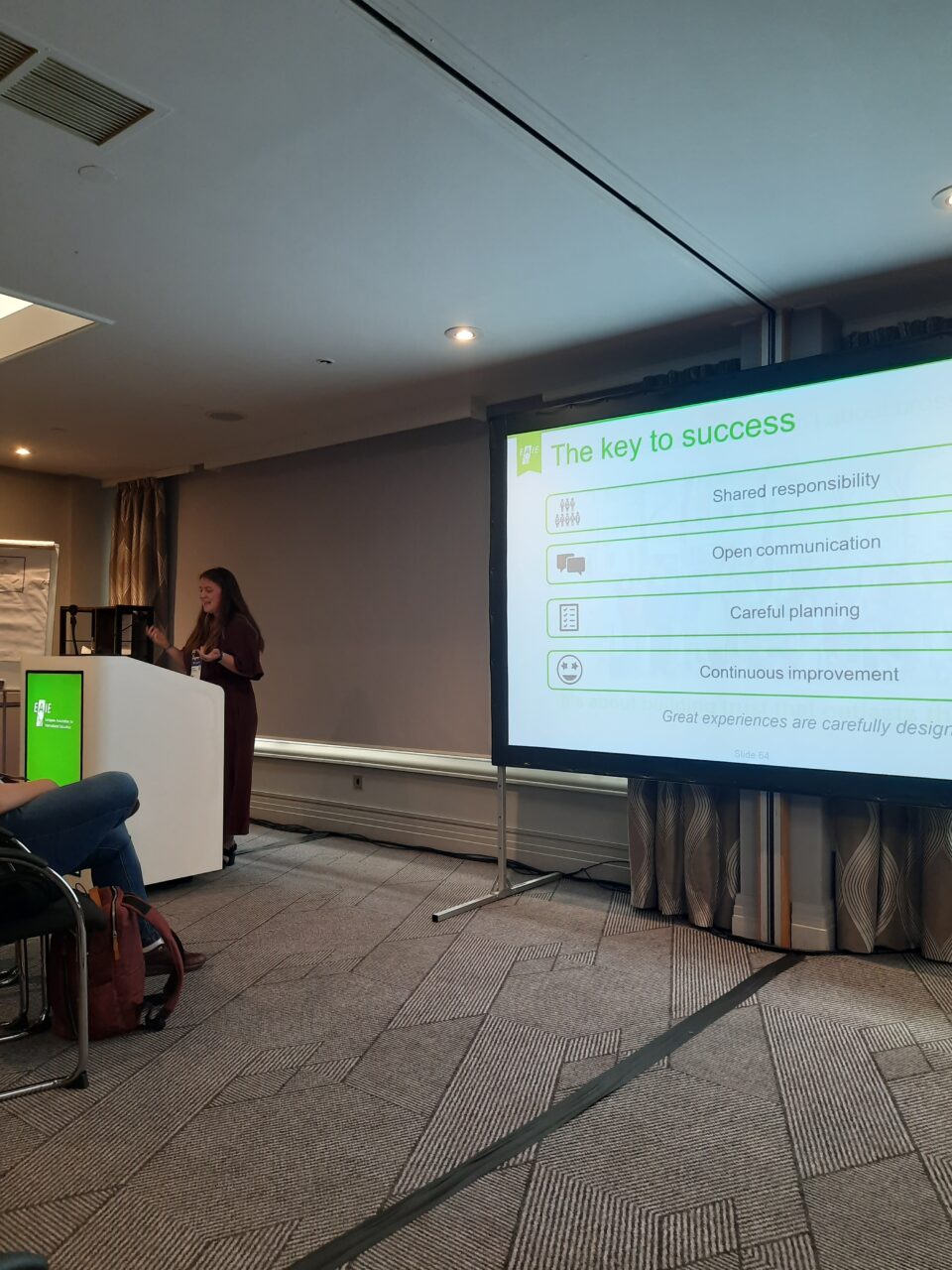 Klára Frausová speaking at a lectern with a green EAIE display, gesturing with both hands. The screen beside her shows a slide titled "The key to success" listing four points: "Shared responsibility", "Open communication", "Careful planning" and "Continuous improvement", followed by the line "Great experiences are carefully designed".