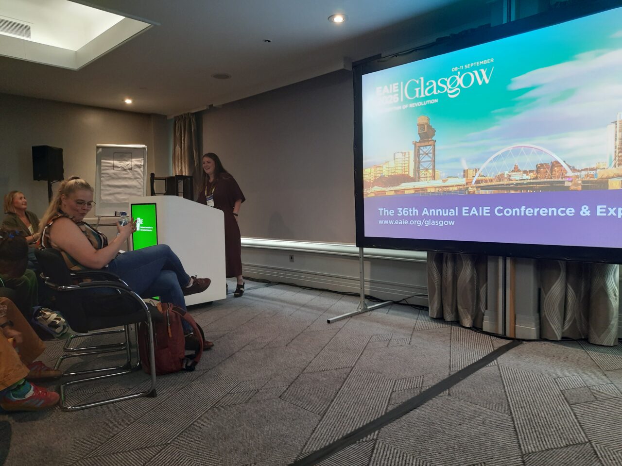 Klára Frausová stands smiling beside a lectern with a green EAIE display at the front of a conference room. Next to her, a large screen shows the EAIE 2026 Glasgow title slide with a view of the city's riverside and the text "The 36th Annual EAIE Conference & Expo". Several audience members are seated in the foreground on the left.
