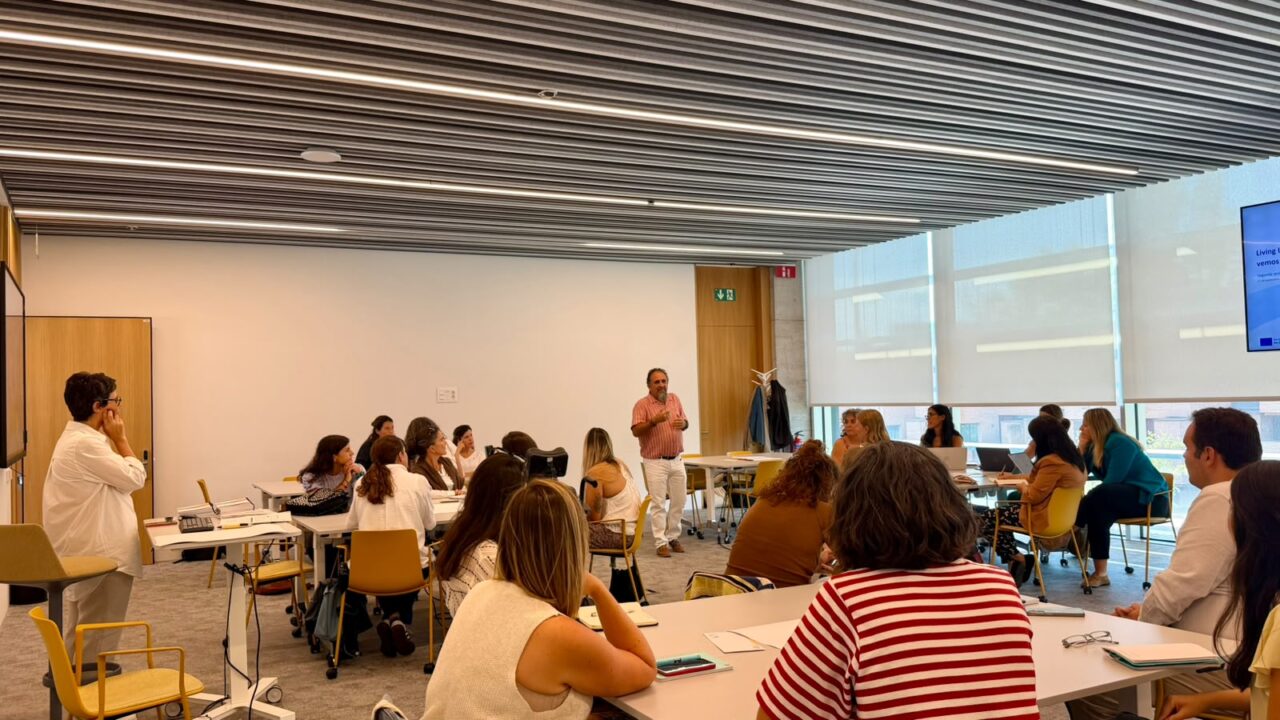 A man stands in the middle of a bright meeting room, speaking to participants seated at tables arranged in a U-shape. A screen on the right shows the title of the Living Lab session.