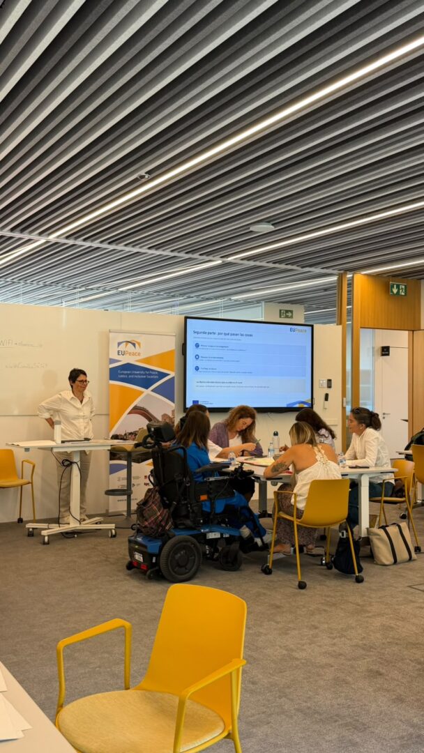 A group of participants, one of them using a power wheelchair, works around a table with papers and markers. A facilitator stands at a high table beside a EUPeace roll-up banner. A screen behind them shows a slide titled "Segunda parte: por qué pasan las cosas".