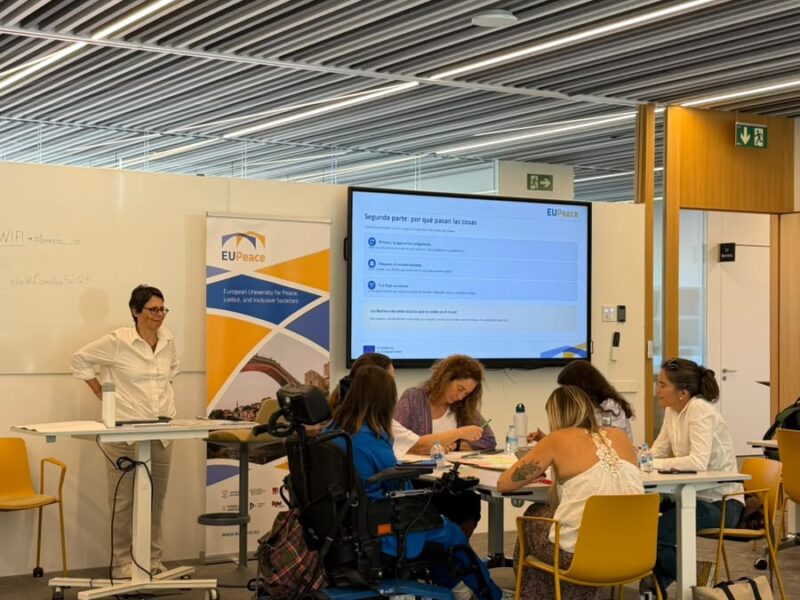 A group of participants, one of them using a power wheelchair, works around a table with papers and markers. A facilitator stands at a high table beside a EUPeace roll-up banner. A screen behind them shows a slide titled "Segunda parte: por qué pasan las cosas".