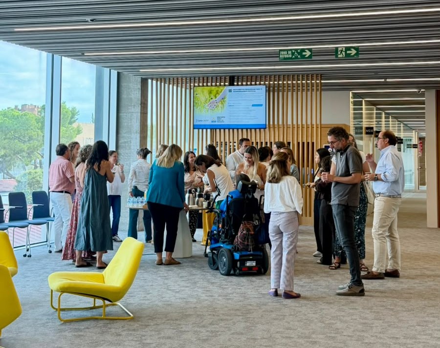 A large group of people, including a participant using a power wheelchair, stands and talks around a coffee table in a spacious, light-filled hall with large windows and yellow armchairs.