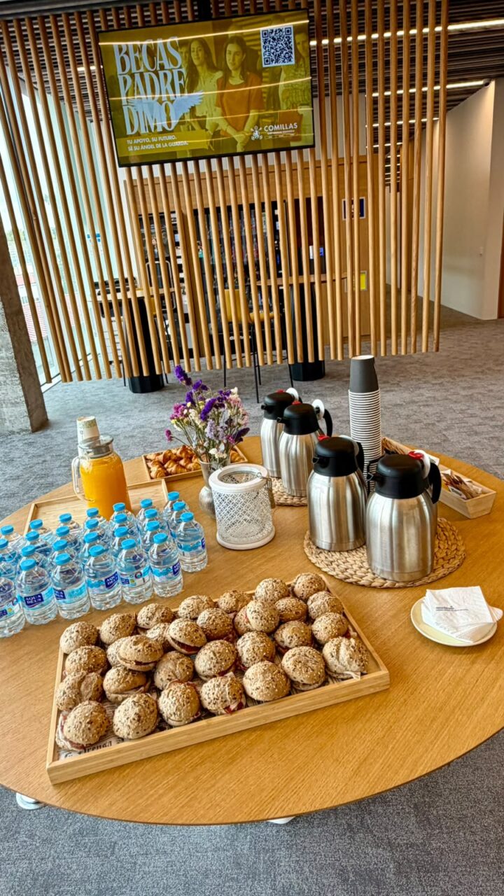 A round wooden table set for a coffee break, with trays of sandwiches and croissants, bottled water, orange juice, coffee thermoses, paper cups and a small vase of flowers. A Comillas University screen hangs in the background.