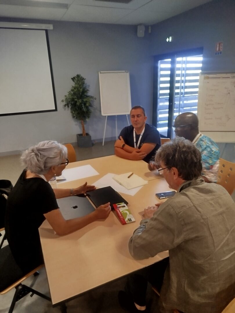 Four people sit around a light wooden table in a meeting room, deep in discussion. A woman with silver hair and glasses gestures towards her laptop while a man in a dark T-shirt listens with folded arms. Two further participants, one in a patterned shirt and one in a grey jacket, lean in over notebooks and papers. A projection screen, a flip chart and a whiteboard with handwritten notes are visible in the background.