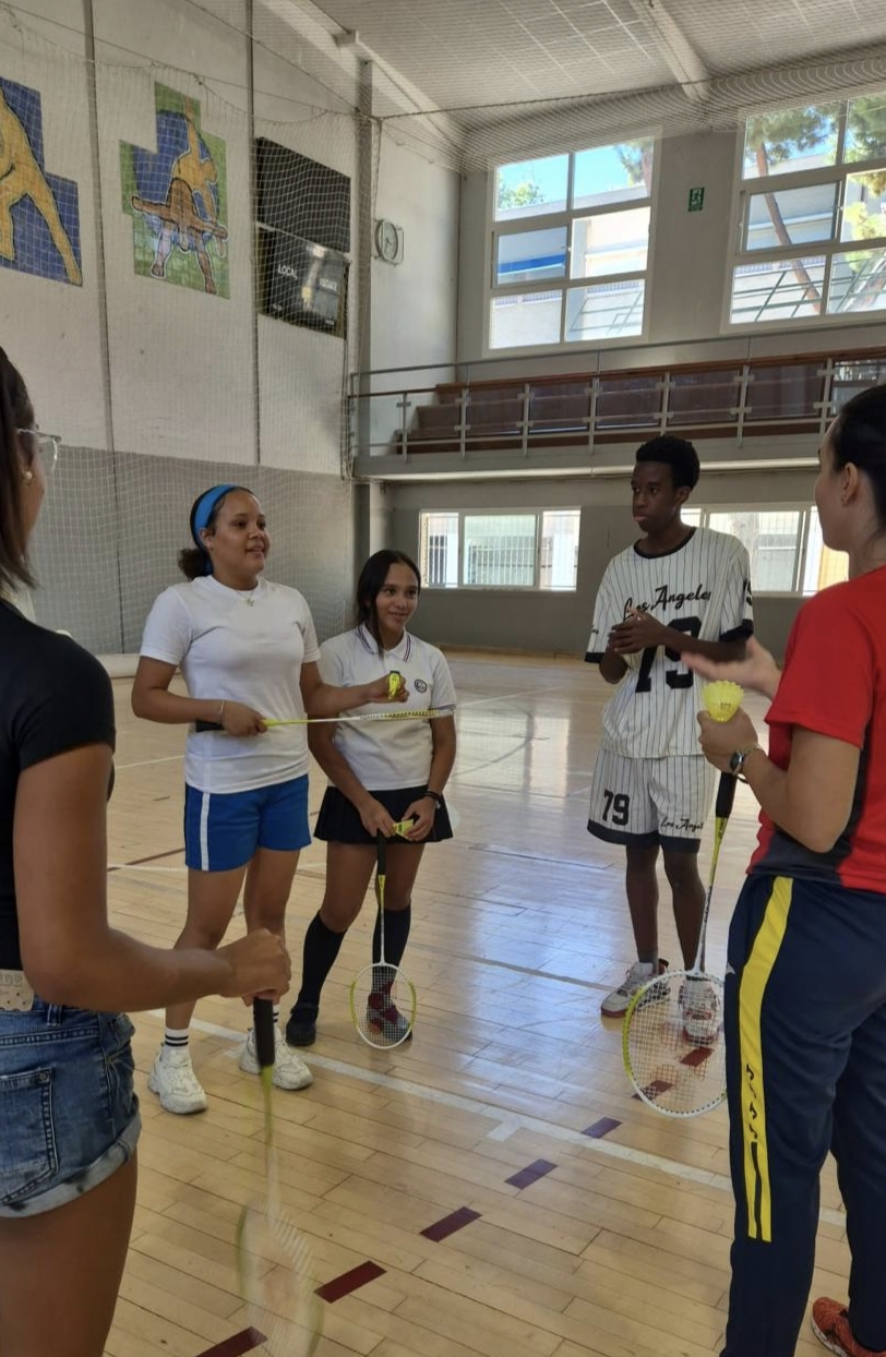 Teenagers holding badminton rackets and shuttlecocks listen to an instructor in an indoor sports hall.