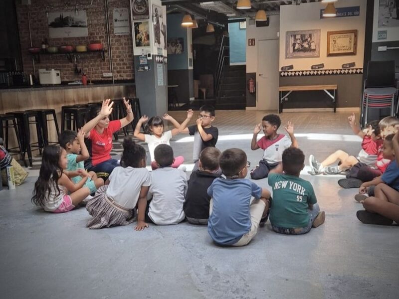 Young children sit in a circle on the floor, copying a leader's raised-hand gestures in an indoor community space.
