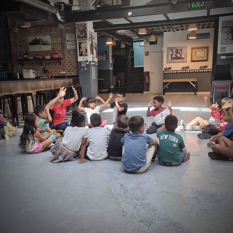 Young children sit in a circle on the floor, copying a leader's raised-hand gestures in an indoor community space.