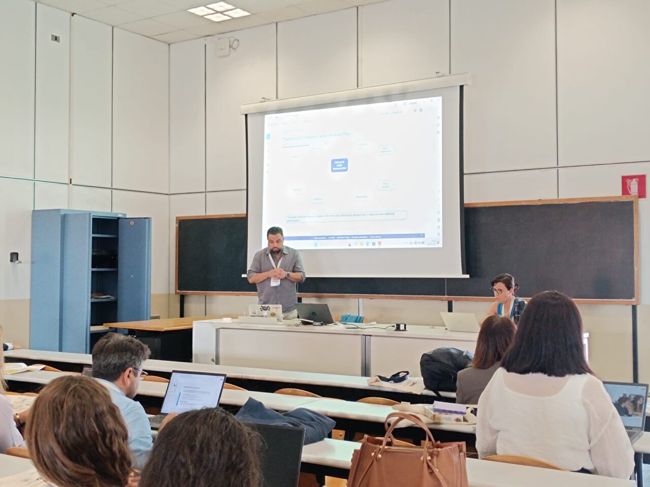 A speaker with a conference badge stands at the front of a classroom and presents to an audience, with a slide on a theoretical framework projected on the screen behind him. A session chair sits at a laptop at the table beside him. Participants in the foreground follow the talk, several with open laptops.