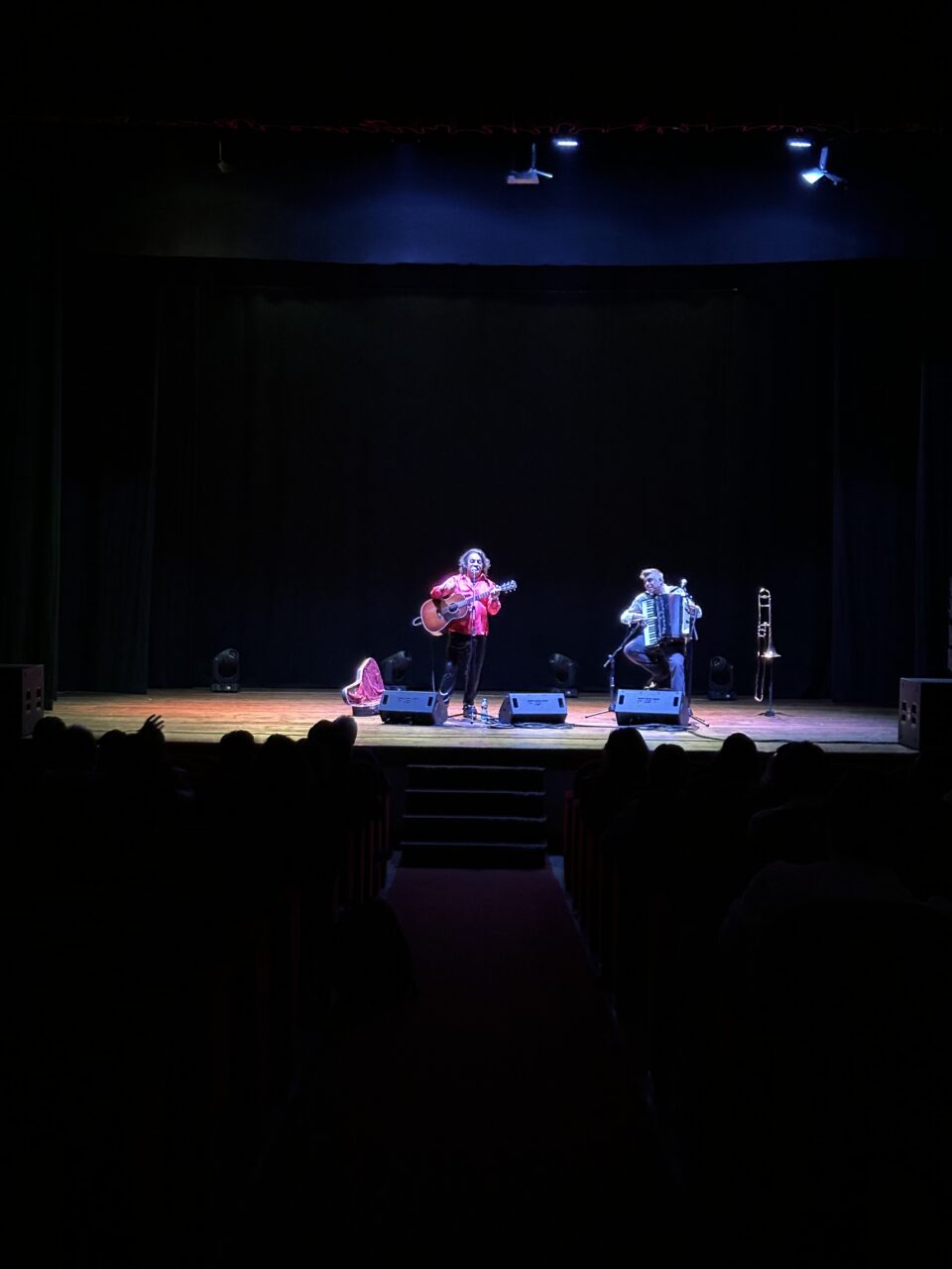 Two musicians perform on a dimly lit theatre stage: on the left, a singer in a red shirt plays an acoustic guitar; on the right, a seated musician plays the accordion, with a trombone on a stand beside him. The silhouettes of the audience are visible in the foreground.