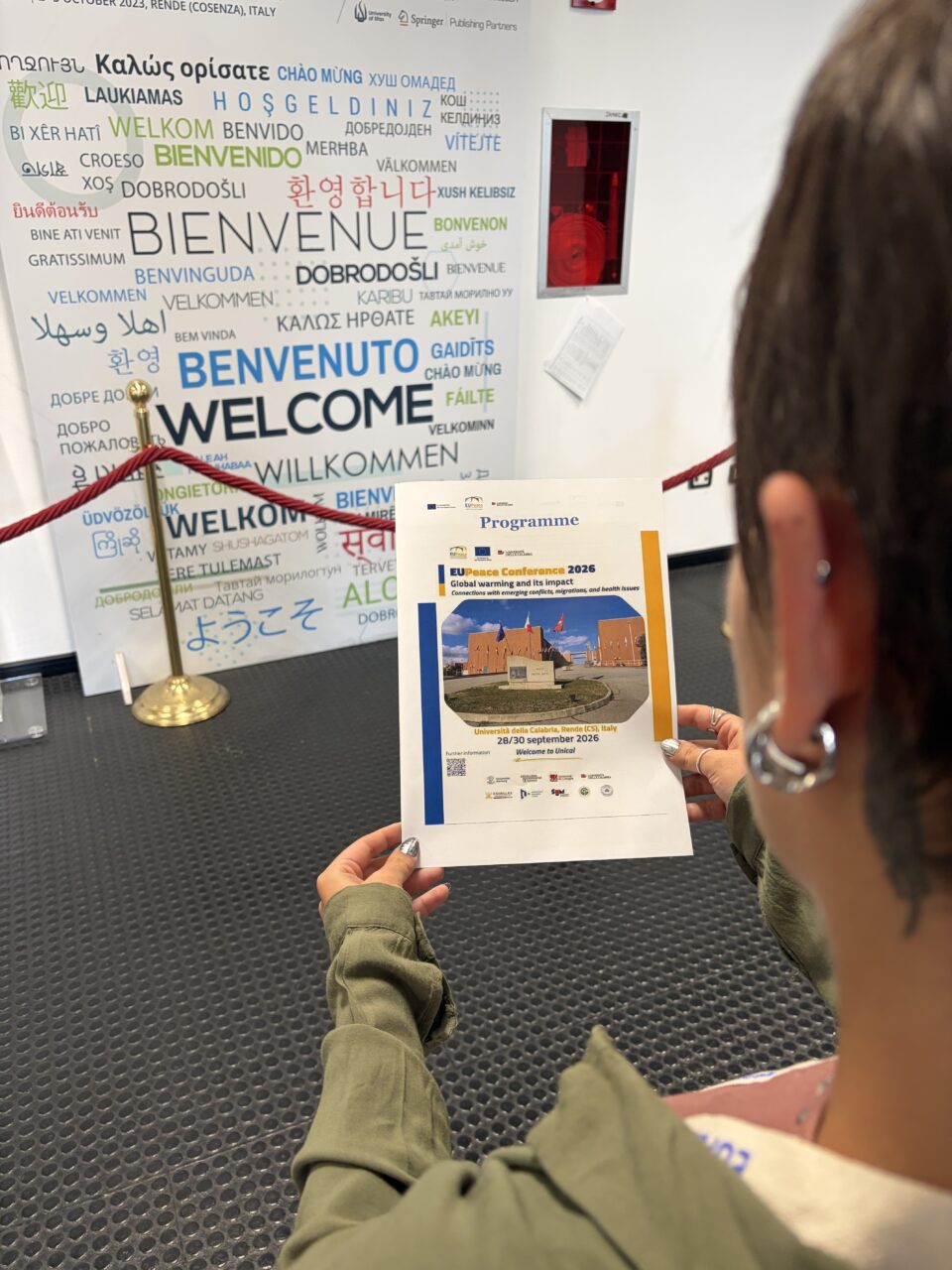 A person, seen from behind, holds the printed programme of the EUPeace Conference 2026. The cover shows the title "Global warming and its impact: Connections with emerging conflicts, migrations, and health issues", a photo of the University of Calabria campus with flags, and the dates 28–30 September 2026. In the background is a large wall with the word "Welcome" in many languages.