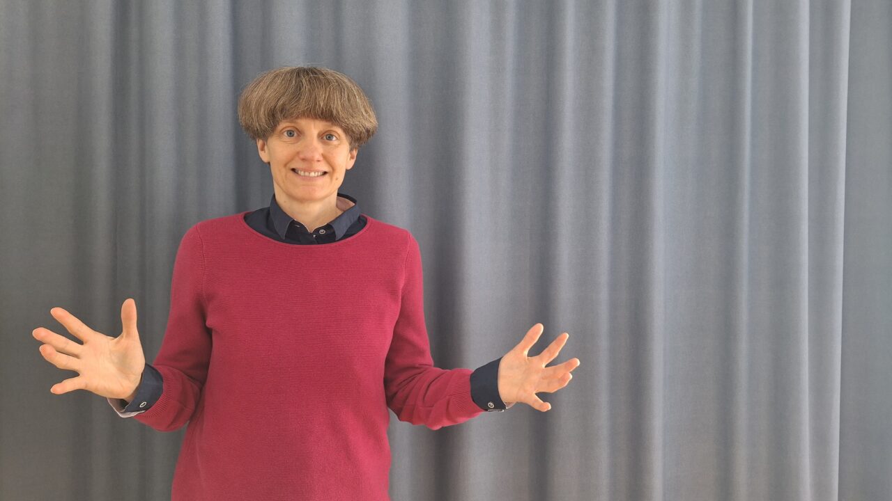 Prof. Edith Braun smiling with both hands open in front of a grey curtain.