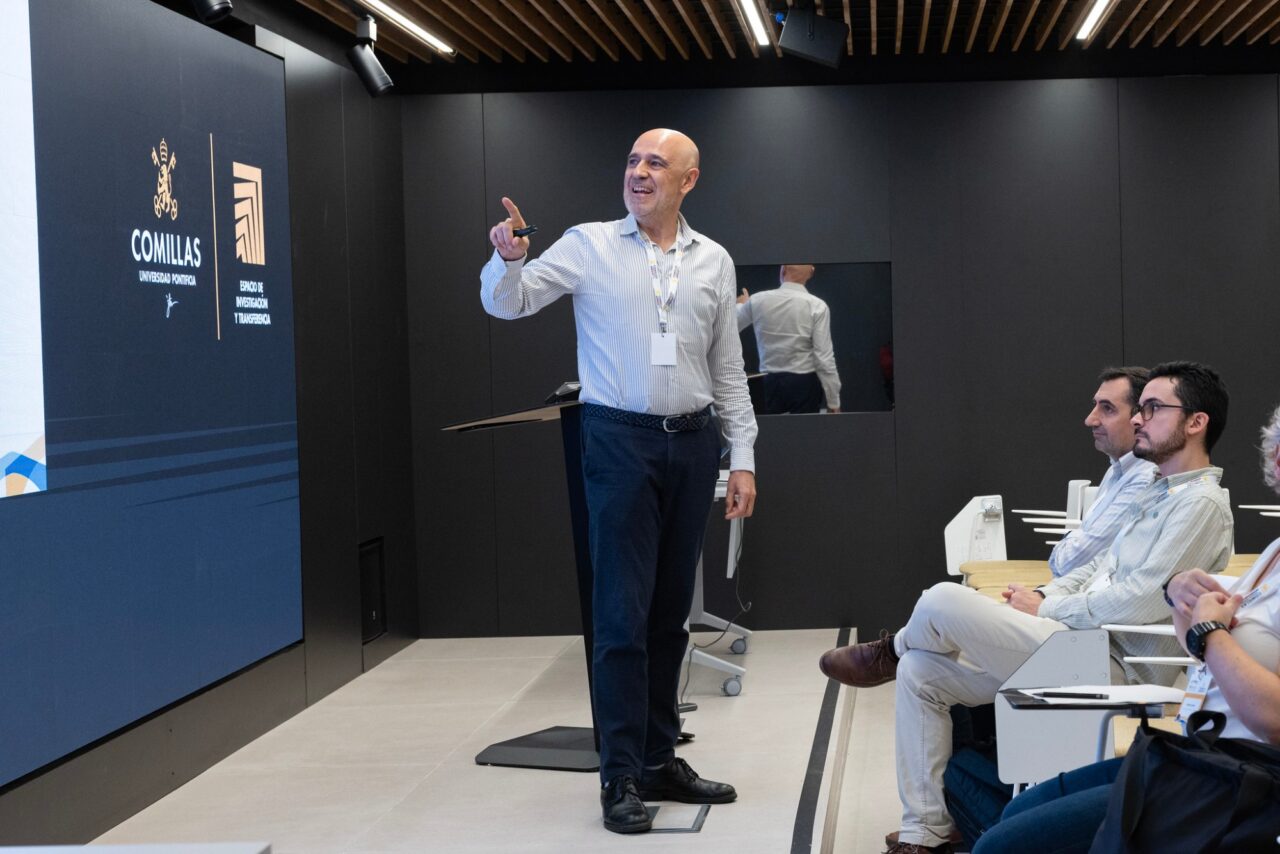 Efraim Centeno, smiling, points towards a large screen showing the logos of Comillas Universidad Pontificia and Espacio de Investigación y Transferencia. On the right, José Carlos Romero and Roberto Barrella sit in the front row with other participants.
