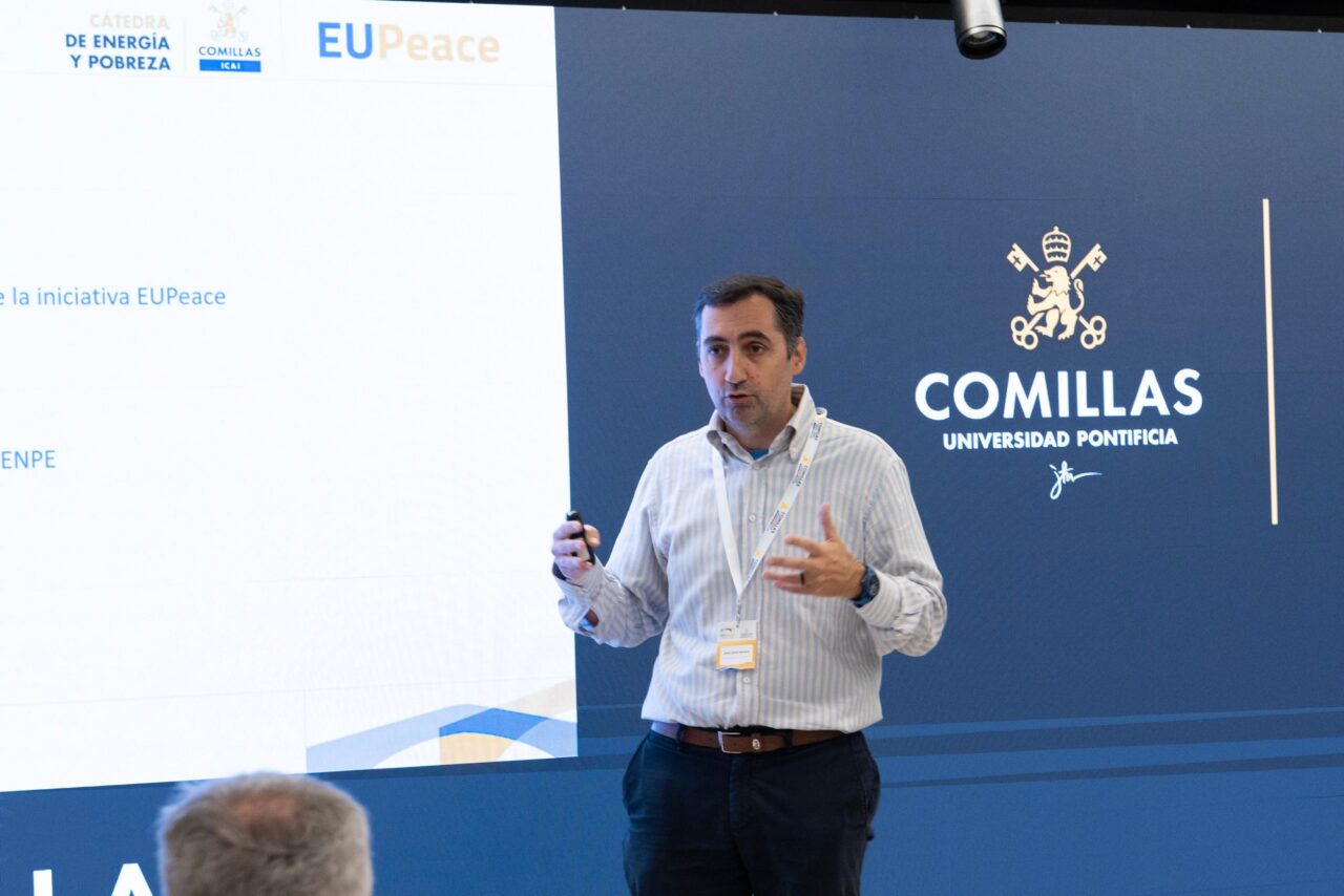 José Carlos Romero, wearing a striped shirt and a conference lanyard, speaks while holding a presentation remote. Behind him, a large screen shows a slide with the logos of the Cátedra de Energía y Pobreza, Comillas ICAI and EUPeace, and the Comillas Universidad Pontificia logo on a dark blue background.