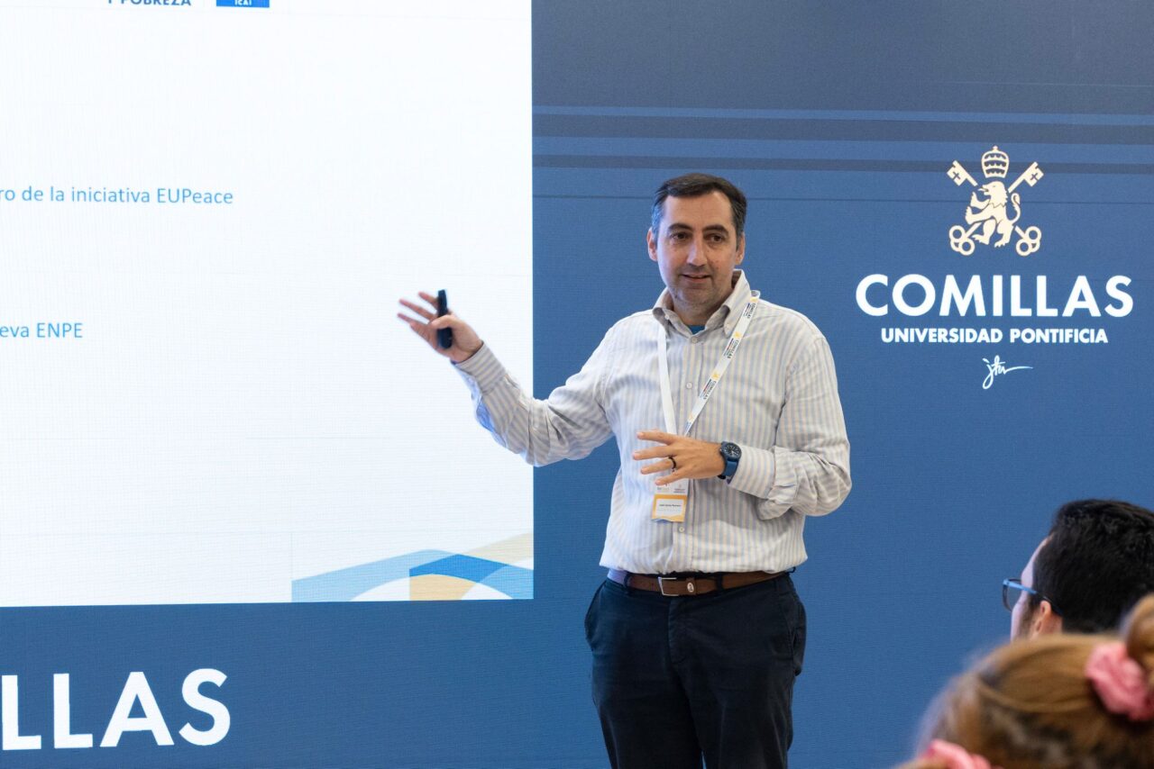 José Carlos Romero, wearing a striped shirt and a conference lanyard, gestures towards a projected slide while holding a presentation remote. The slide partly shows the words “la iniciativa EUPeace” and “ENPE”. The Comillas Universidad Pontificia logo is on the right, and two audience members are in the foreground.