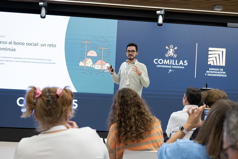 Roberto Barrella, wearing glasses, a light shirt and a lanyard, presents in front of a screen. The slide reads “El acceso al bono social: un reto que continúa” next to an illustration of houses and an electricity pylon, beside the logos of Comillas Universidad Pontificia and Espacio de Investigación y Transferencia. Audience members are seen from behind in the foreground.