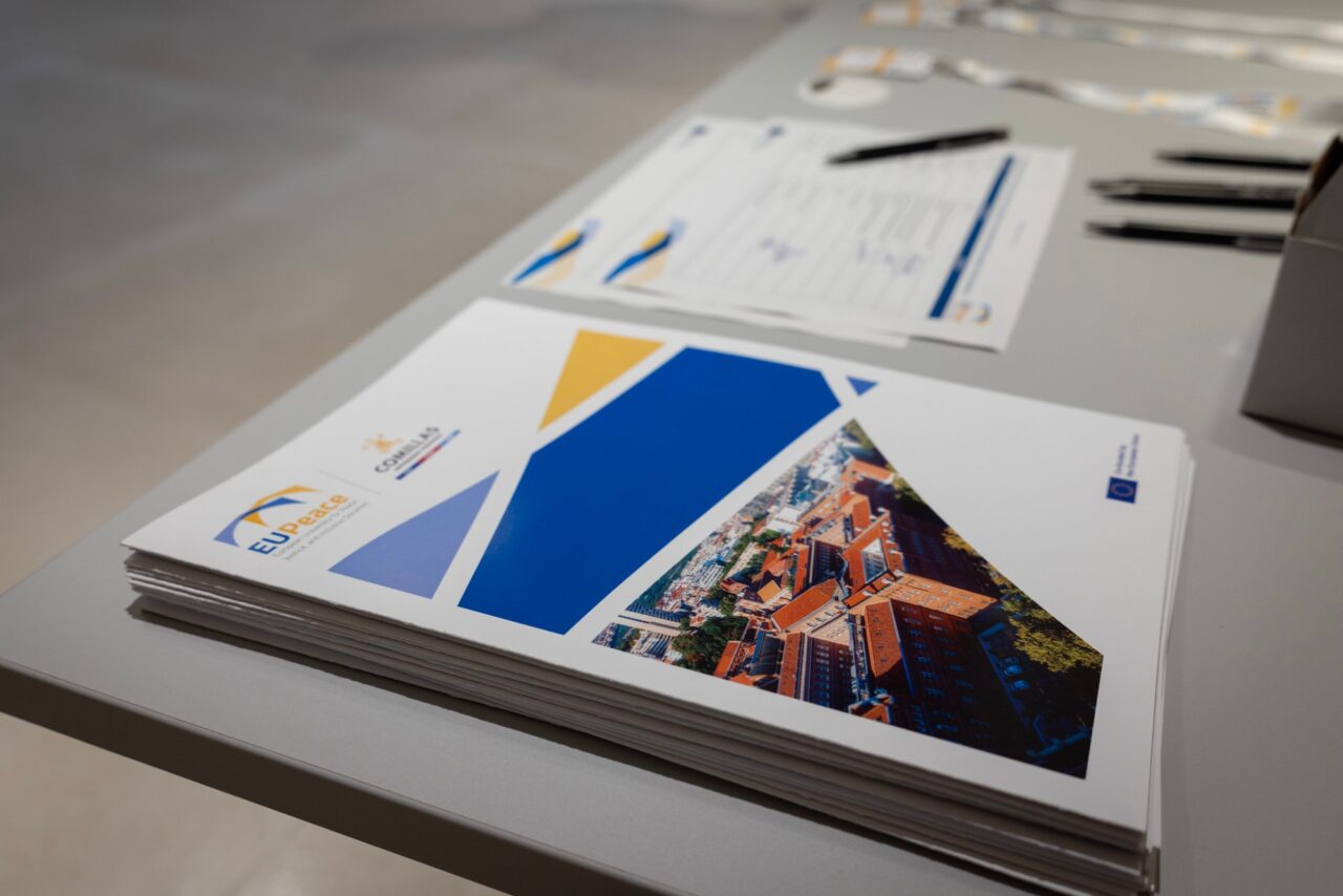 A stack of folders on a grey table. The folders show the EUPeace and Comillas logos, blue and yellow geometric shapes and an aerial photo of red-roofed university buildings. Sign-in sheets, pens and conference lanyards lie behind them.