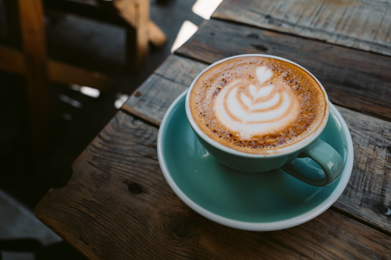 A cappuccino with a leaf-shaped latte art pattern in a turquoise cup and matching saucer, standing on a rustic wooden table.