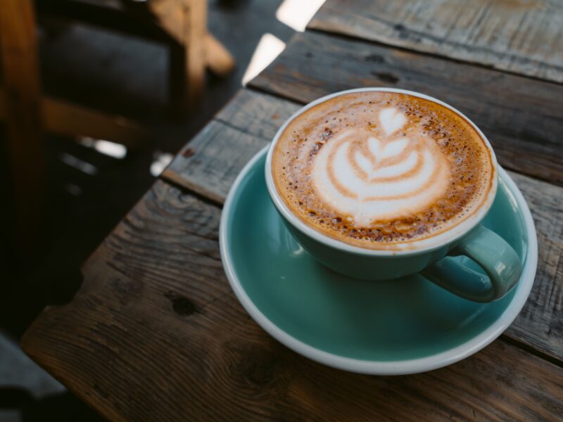 A cappuccino with a leaf-shaped latte art pattern in a turquoise cup and matching saucer, standing on a rustic wooden table.