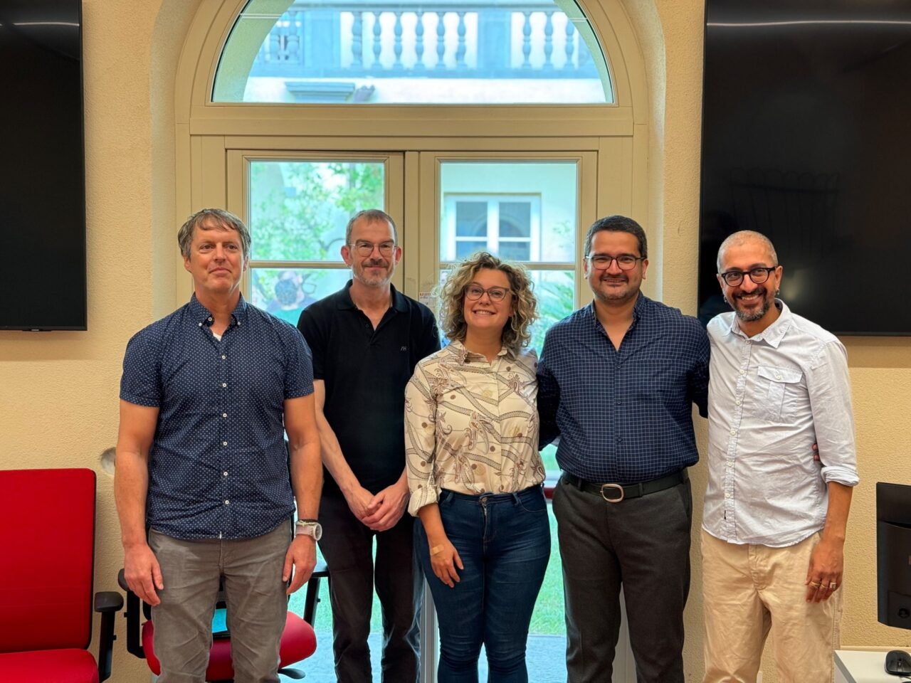 Five people stand side by side in a row, smiling at the camera, in a room with cream-coloured walls. Four men and one woman with curly hair are wearing casual clothes, and some of them wear glasses. Behind them is a tall arched window overlooking greenery and a building, with dark wall-mounted screens on either side and a red office chair at the left.