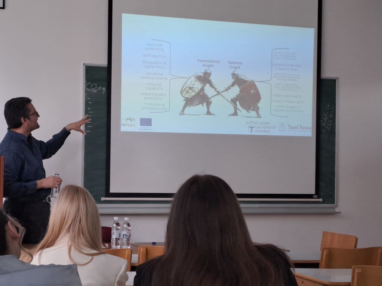 A man in a checked shirt and glasses stands beside a projector screen in a classroom, holding a water bottle and gesturing towards a slide. The slide shows two armoured knights fighting, labelled "Transnational knight" and "National knight", with lists of terms on either side such as free trade agreements, tariff reduction, weaker public welfare and erosion of labour rights. Two seated audience members are seen from behind in the foreground.
