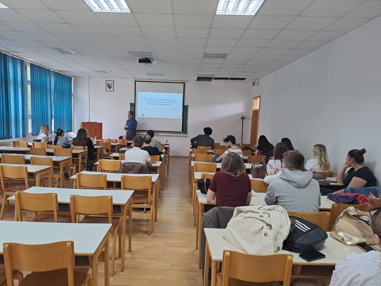A man stands at the front of a classroom beside a projection screen, giving a presentation to a small audience seated at wooden desks. The slide reads "The Creation of a Neoliberal Autarky: When Does Developmentalism Become Anti-Development?"