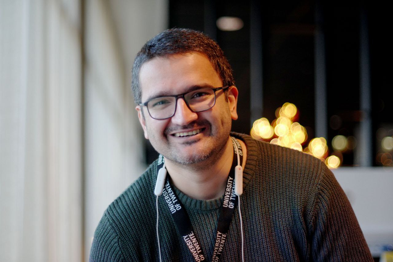 A smiling man with short dark hair, a stubbled beard and dark-framed glasses, wearing a dark green knitted jumper. A black University of Twente lanyard and white wireless earphones hang around his neck. Blurred warm lights are visible behind him.