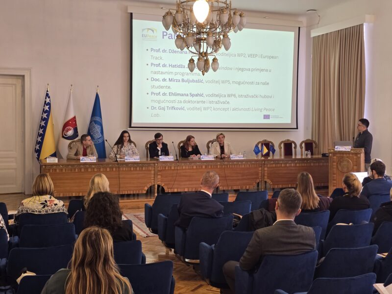 A panel of five people sits at a long wooden table in a formal hall with a chandelier, while a person stands at a lectern to the right. Behind them a projected slide carries the EUPeace logo and a Bosnian-language list of names and roles, including work package leads and a Living Peace Lab activity. Three flags stand to the left, and audience members sit with their backs to the camera in blue chairs.