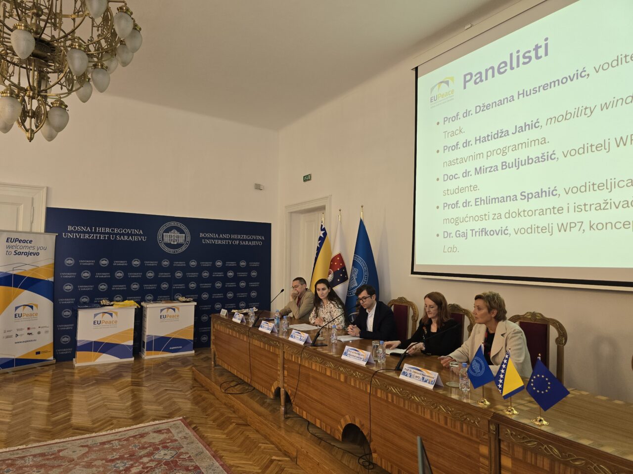 Five panellists sit behind a long wooden table in a formal hall with a chandelier, parquet floor and a blue University of Sarajevo backdrop. A projected slide titled "Panelisti" lists speakers, and EUPeace banners and small flags stand nearby.