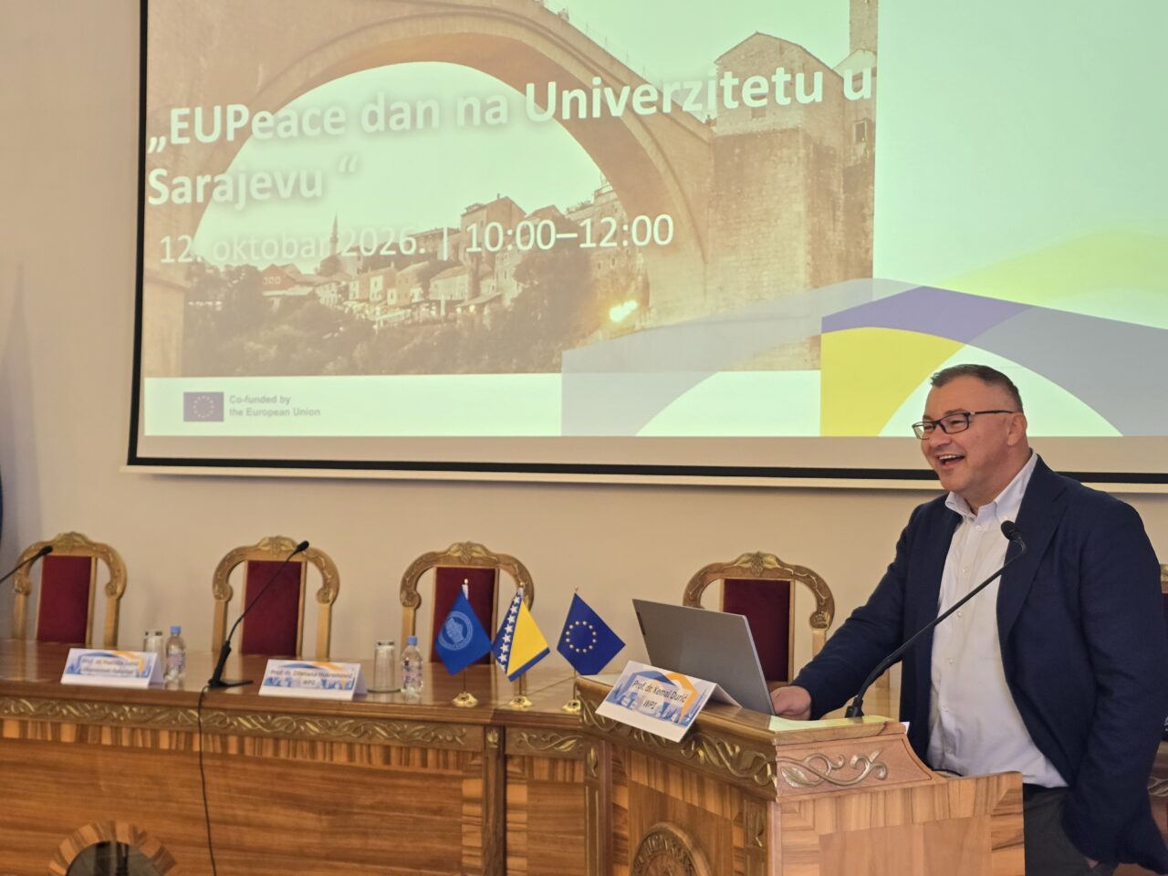 A smiling man with glasses, wearing a dark blazer and light shirt, speaks at a wooden lectern with a laptop and microphone. Behind him, a projected slide reads "EUPeace dan na Univerzitetu u Sarajevu", 12 October 2026, 10:00–12:00, with a stone bridge image and the text "Co-funded by the European Union".
