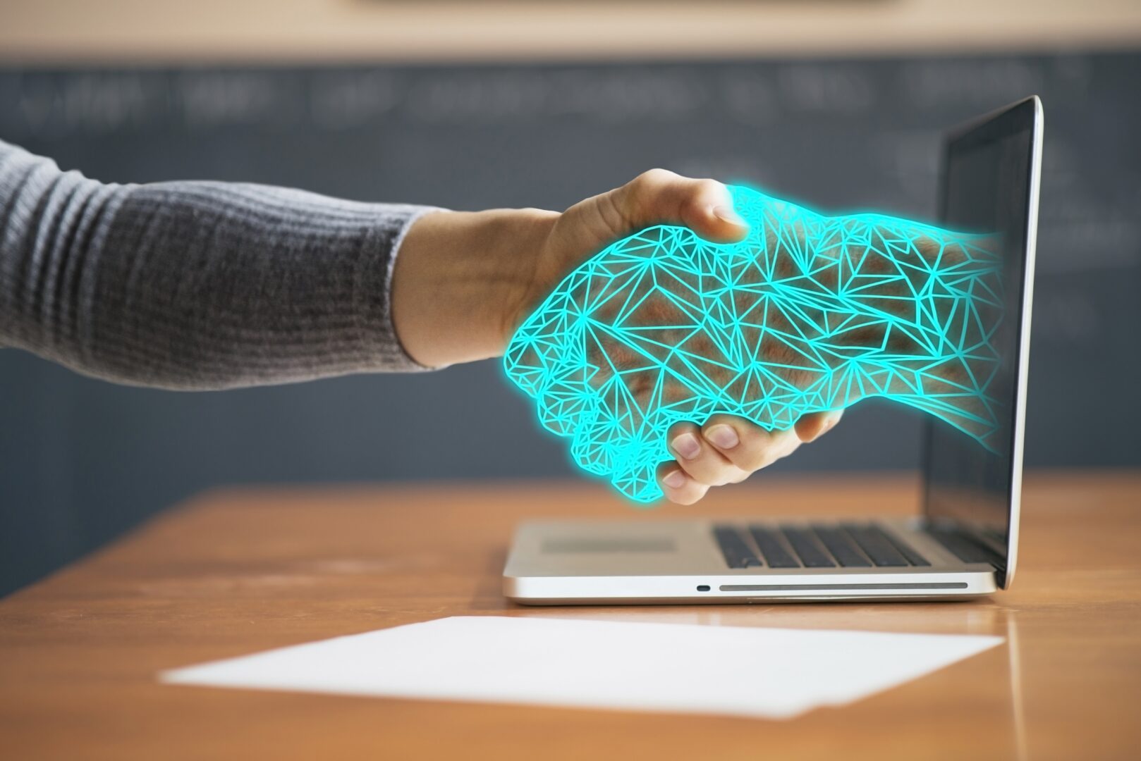A human hand in a grey sleeve shakes a glowing, turquoise wireframe hand that extends from the screen of an open laptop. The laptop stands on a wooden desk next to a blank sheet of paper, with a blurred blackboard in the background.