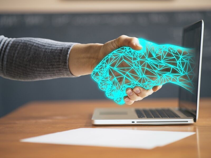 A human hand in a grey sleeve shakes a glowing, turquoise wireframe hand that extends from the screen of an open laptop. The laptop stands on a wooden desk next to a blank sheet of paper, with a blurred blackboard in the background.