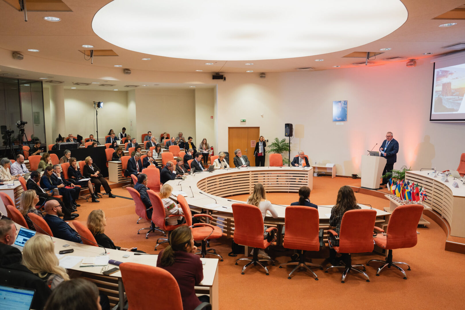 Participants seated around curved conference tables listen to a speaker at a lectern during the Magna Charta gathering in Vilnius.