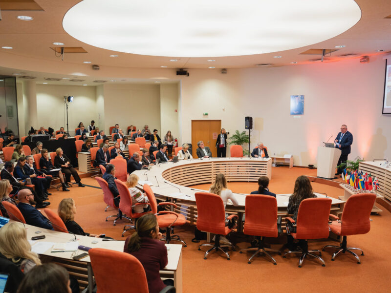 Participants seated around curved conference tables listen to a speaker at a lectern during the Magna Charta gathering in Vilnius.