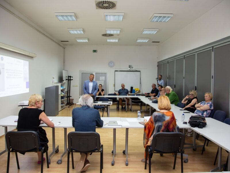 A doctoral dissertation defence in a seminar room at the Centre for Interdisciplinary Studies, University of Sarajevo. A man in a light blue jacket stands at the back of the room next to a projected slide reading 'Doktorska disertacija' and the title 'Procjena kvalitete usluga bosanskohercegovačkog visokog obrazovanja – primjena SERVQUAL modela'. Three committee members are seen from behind, seated at tables in the foreground, and members of the audience sit along the tables on the right.