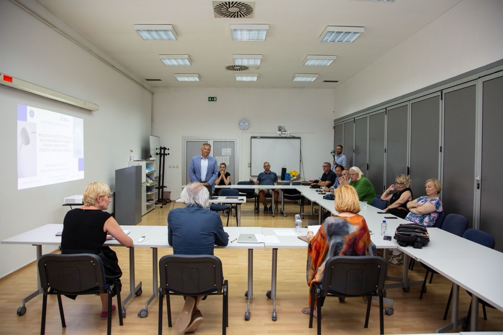 A doctoral dissertation defence in a seminar room at the Centre for Interdisciplinary Studies, University of Sarajevo. A man in a light blue jacket stands at the back of the room next to a projected slide reading 'Doktorska disertacija' and the title 'Procjena kvalitete usluga bosanskohercegovačkog visokog obrazovanja – primjena SERVQUAL modela'. Three committee members are seen from behind, seated at tables in the foreground, and members of the audience sit along the tables on the right.
