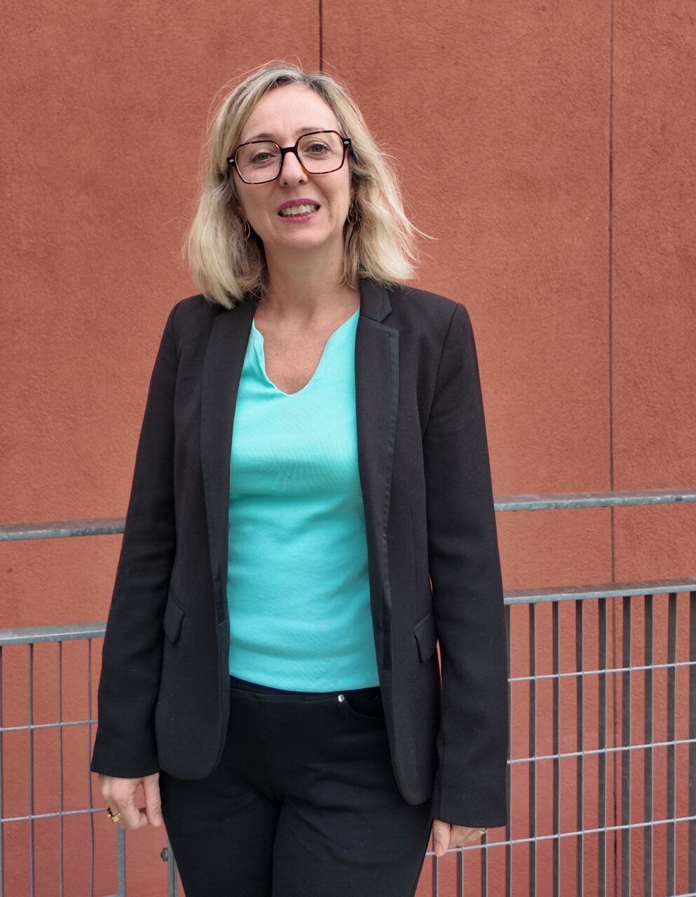 Caroline Foulquier stands outdoors, wearing dark-framed glasses and a black blazer over a turquoise top, in front of a terracotta-coloured wall.