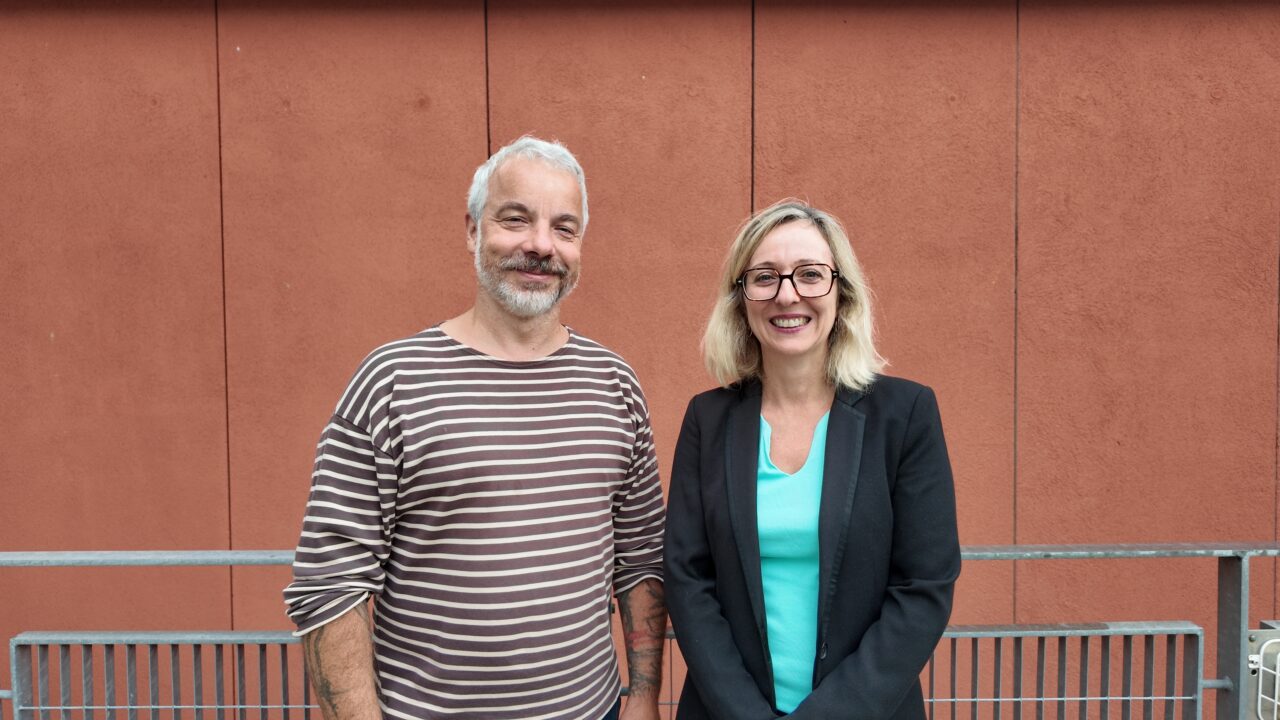 Petr Šimon and Caroline Foulquier stand side by side, smiling, in front of a terracotta-coloured panelled wall.