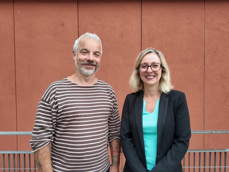 Petr Šimon and Caroline Foulquier stand side by side, smiling, in front of a terracotta-coloured panelled wall.