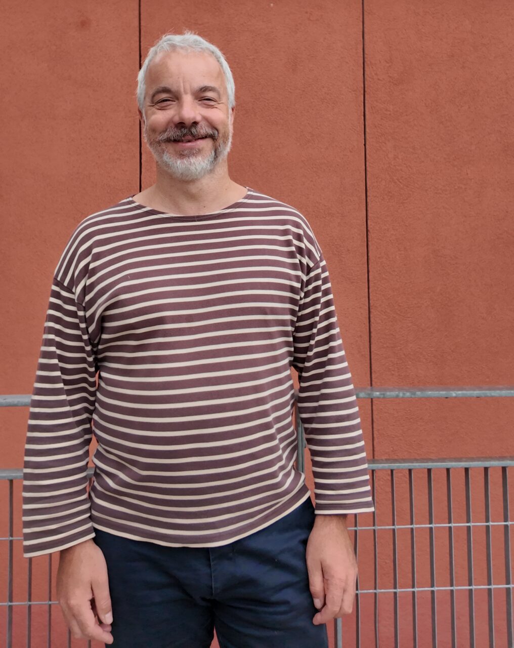 Petr Šimon stands outdoors, smiling, in a brown-and-white striped long-sleeved top in front of a terracotta-coloured wall.