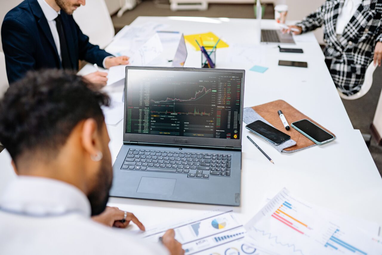 Three people sit around a white table in an office or meeting room. In the foreground a man with dark hair looks at a laptop showing a trading platform with a price chart and order book, while he rests a hand on printed charts. Two other people, one in a dark suit and one in a black-and-white checked jacket, work with papers and a laptop in the background.