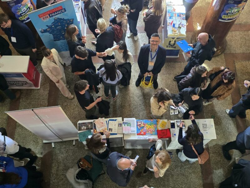 Aerial view of a crowded exhibition hall with stands and students, one man in the centre smiling up at the camera.