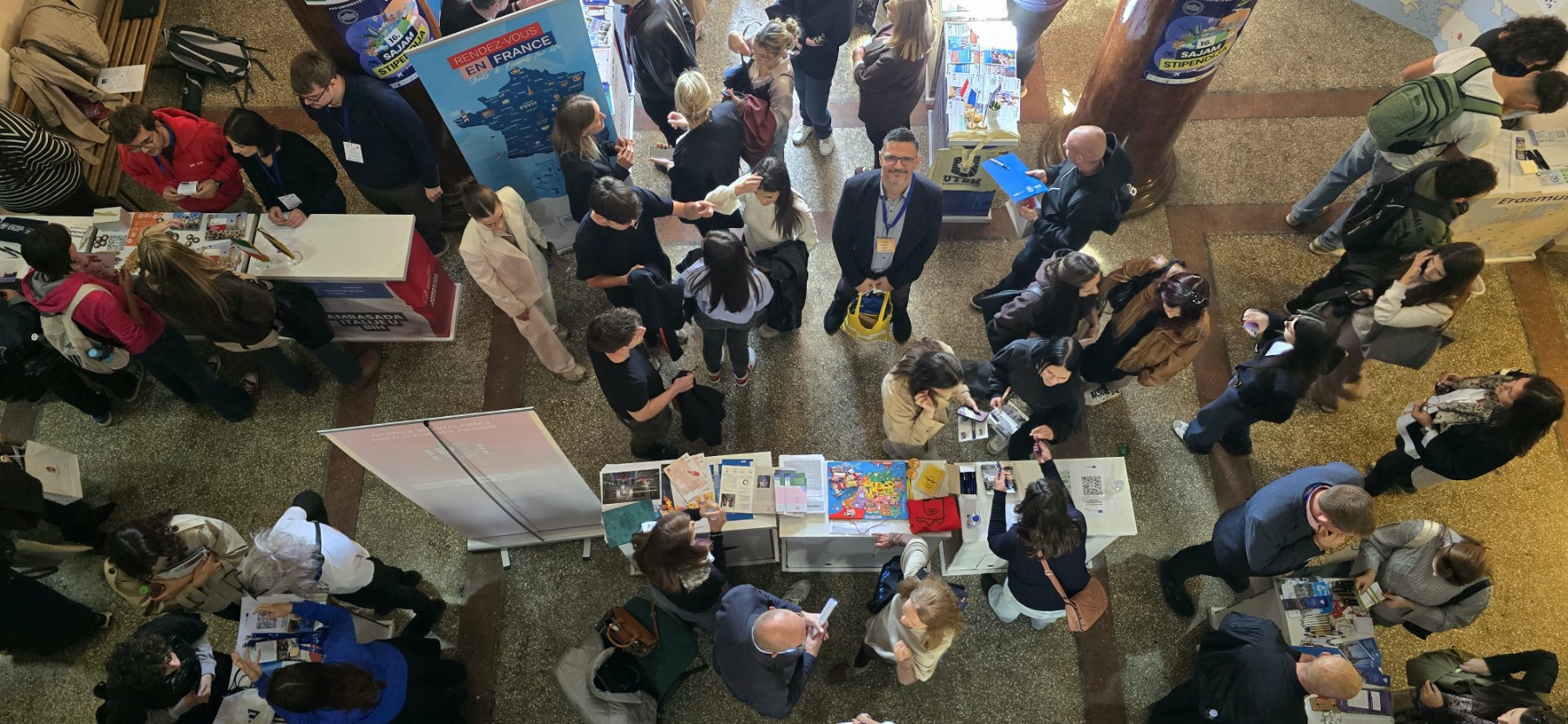 Aerial view of a crowded exhibition hall with stands and students, one man in the centre smiling up at the camera.