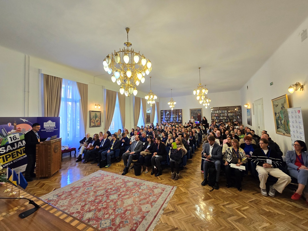 A speaker at a wooden lectern addresses a full hall under crystal chandeliers during the Expo’s opening ceremony.