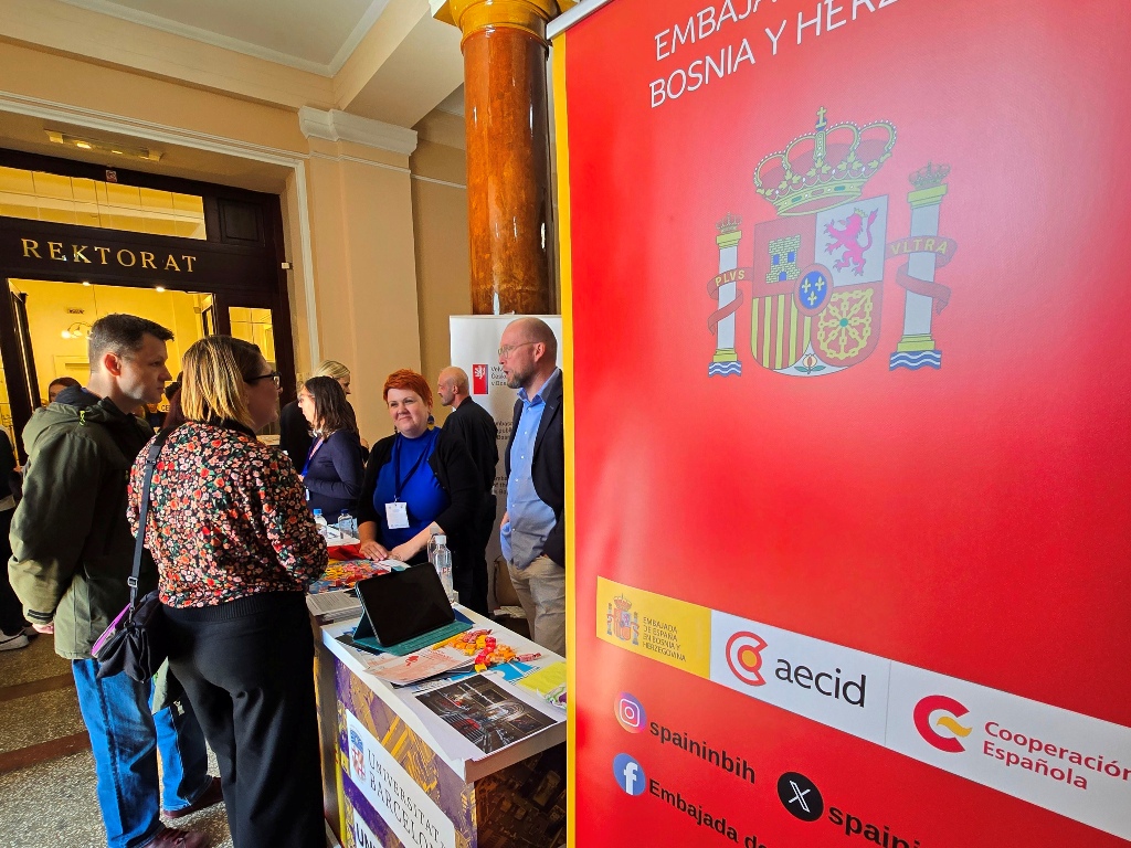 Visitors talk with representatives at the Universitat de Barcelona stand beside a red banner of the Spanish Embassy.