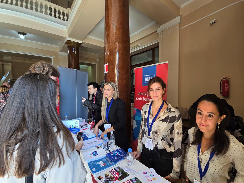 Three women with lanyards stand behind a table of brochures, talking to visitors, next to an Institut français roll-up banner.