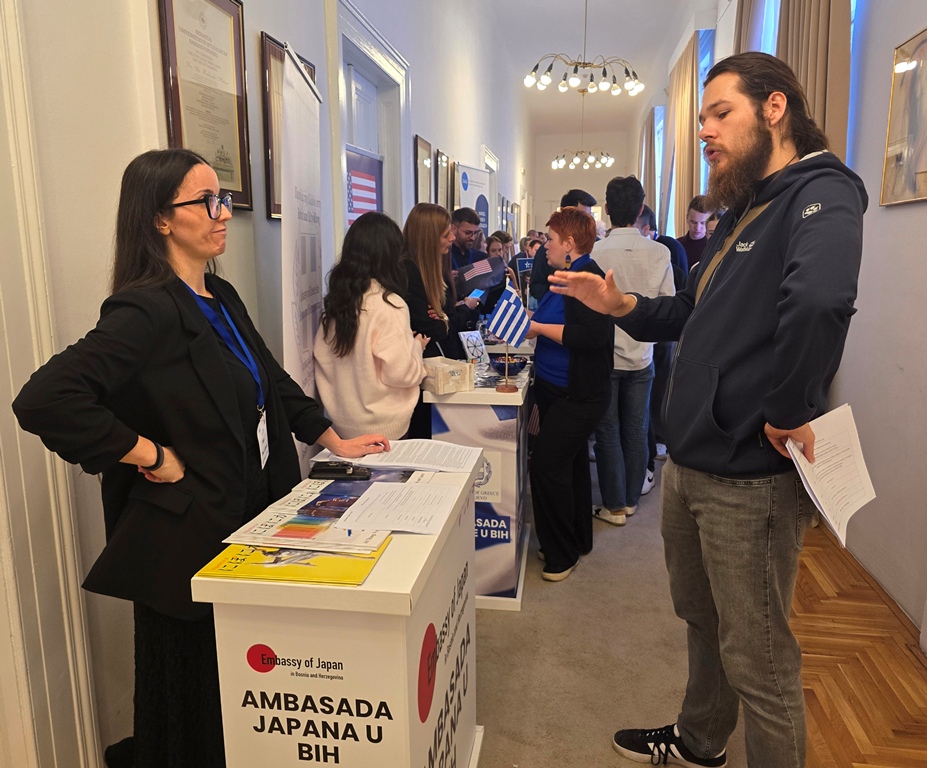 A young man gestures while talking to a representative at the Embassy of Japan stand in a busy corridor lined with stands.